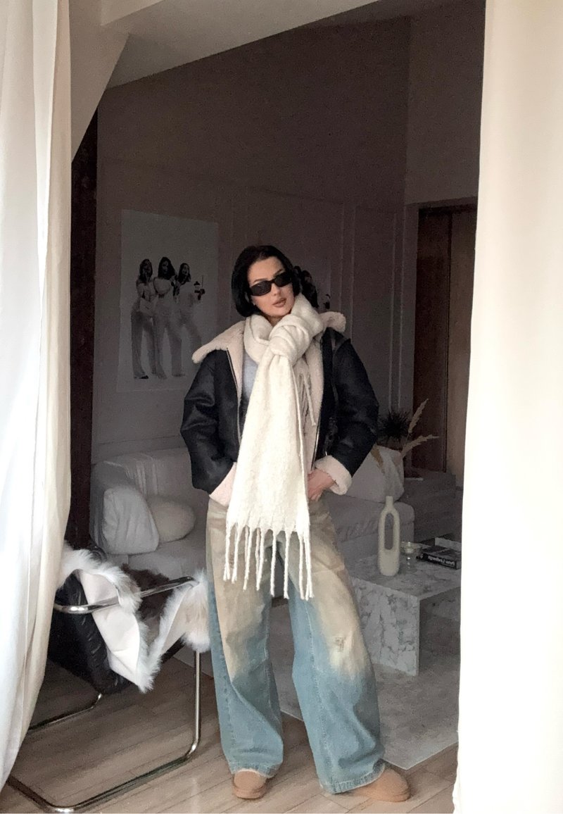 Schwarze Kunstlederjacke mit Schaffellkragen, großer cremefarbener Schal mit Fransen, hellblaue weite Jeans und beigen Slippern.