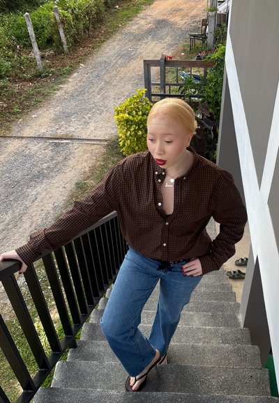 Persona de piel clara y cabello rubio, vestida con una camisa marrón a cuadros y jeans azules, de pie en unas escaleras al aire libre sujetando la barandilla.