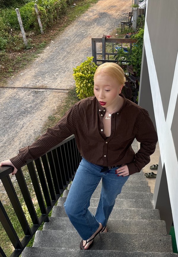 Personne à la peau claire et aux cheveux blonds portant une chemise marron à carreaux et un jean bleu, debout sur des escaliers extérieurs en tenant la rampe.