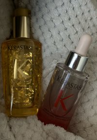 Kérastase matu kopšanas produkti: zeltains, teksturēts pumpas pudelīte ar caurspīdīgu vāciņu, uz kura ir uzraksts "Elixir Ultime", un rozā serums pilinātāja pudelīte.