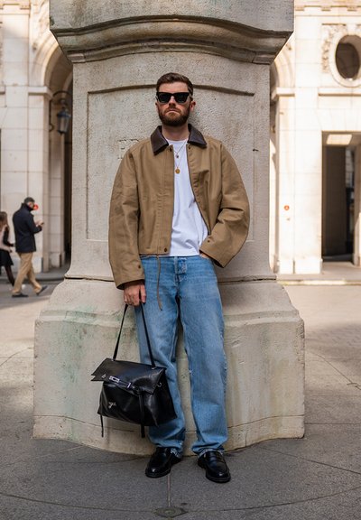 Hombre con barba y gafas de sol de pie junto a un pilar de piedra, vistiendo chaqueta marrón, camisa blanca, vaqueros azules, zapatos negros y sosteniendo una bolsa de cuero negra.