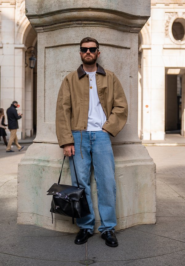 Man with beard and sunglasses stands against a stone pillar, wearing a brown jacket, white shirt, blue jeans, black shoes, and holding a black leather bag.