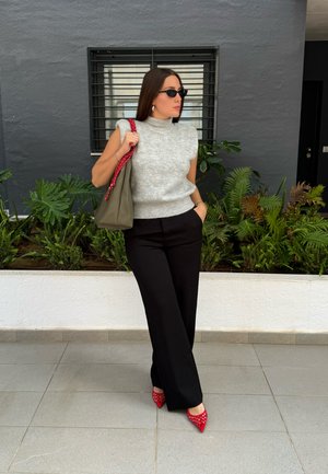 Suéter gris sin mangas, pantalones anchos negros, tacones rojos con tachuelas y un bolso verde oliva con una correa roja, todo sobre un fondo neutro.