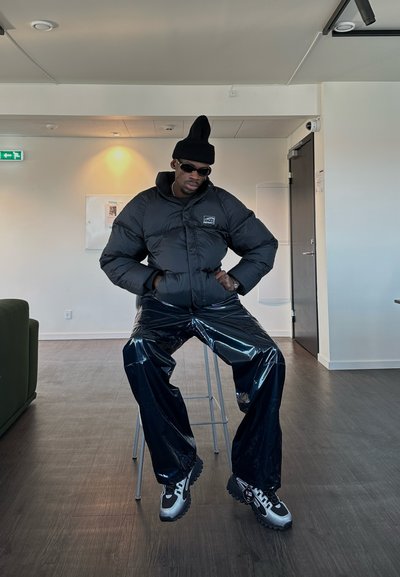 Chaqueta de plumas negra con un logo, pantalones de vinilo negro brillantes, gorro negro y gafas de sol. Zapatillas altas con detalles en gris y negro.