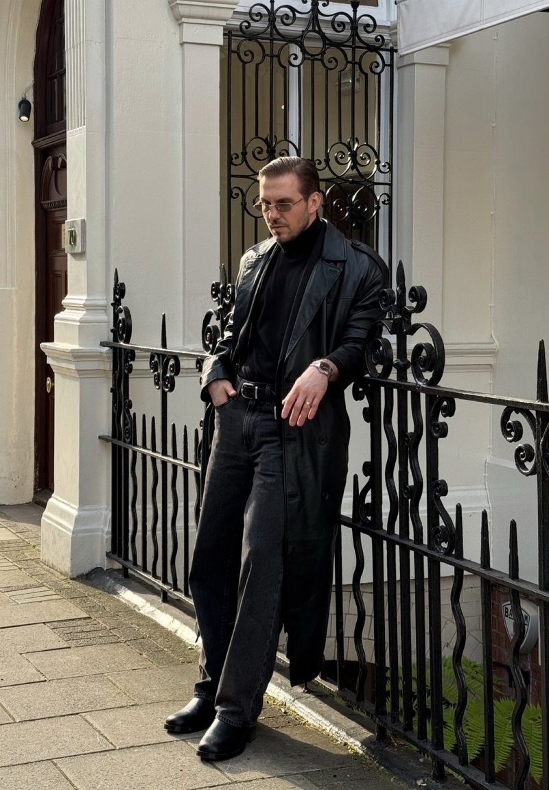 Homme portant un trench-coat en cuir noir, un col roulé noir, des lunettes de soleil et des bottes, appuyé contre une clôture en fer noir ornée sur le trottoir de la ville.