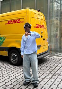 Person som har på seg en lyseblå knappskjorte og store lyse denimjeans, holder en drink, står foran en gul DHL-lastebil.