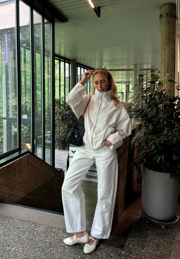 Vrouw in geheel witte outfit en witte schoenen staat binnen bij grote ramen en planten, ze zet haar zonnebril met één hand recht.
