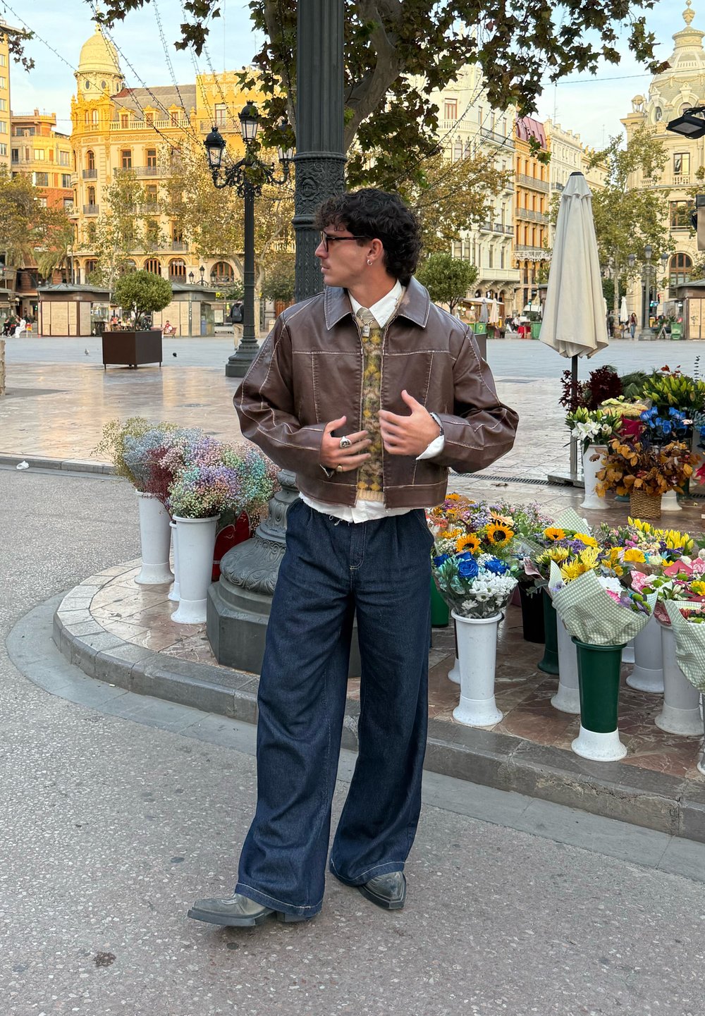 Giacca corta in pelle marrone, camicia a fantasia, pantaloni larghi in denim scuro, scarpe grigie. Circondato da bancarelle di fiori in un ambiente urbano.