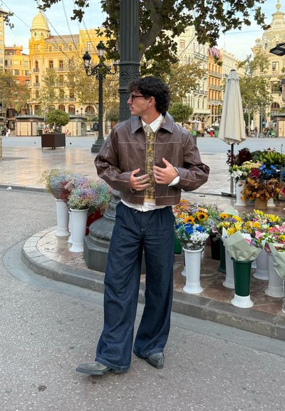 Chaqueta corta de cuero marrón, camisa con estampado, pantalones holgados de mezclilla oscura, zapatos grises. Rodeado de puestos de flores en un entorno urbano.