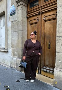 Brūns adīts kardigans virs atbilstošiem platu kāju biksēm, kopā ar melnu somiņu. Apavi sportiska tīrība papildina izskatu pie reljefa akmens sienas.