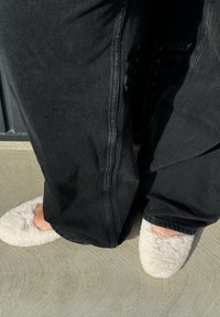 Weiße, flauschige Hausschuhe mit runder Form und Lavendel-Akzenten, getragen mit schwarzen weiten Hosen. Die Oberfläche ist strukturiert und weich.