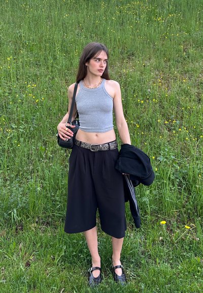 Top corto gris, pantalones de pierna ancha negros con un cinturón de tachuelas y bolso negro. La modelo está de pie sobre la hierba con flores amarillas.
