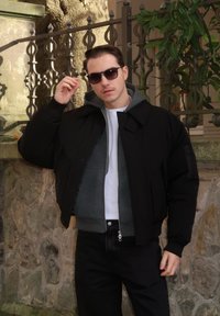 Doudoune noire avec un col montant portée sur un sweat à capuche gris et un t-shirt blanc, associée à un jean foncé et des lunettes de soleil noires. Fond en mur de pierre.