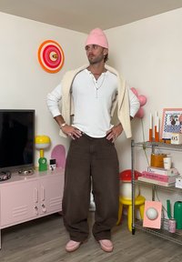 Bruine wijde broek, wit langslavende shirt, beige trui over de schouders, roze beanie en roze schoenen. Kleurrijke decoratie op de achtergrond.