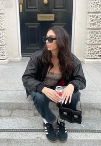 Μαύρο bomber jacket, τοπ με λεοπάρ σχέδιο, μπλε τζιν παντελόνι, μαύρα αθλητικά παπούτσια με λευκές ρίγες, κρατώντας ένα αναψυκτικό, καθισμένο σε πέτρινα σκαλιά.