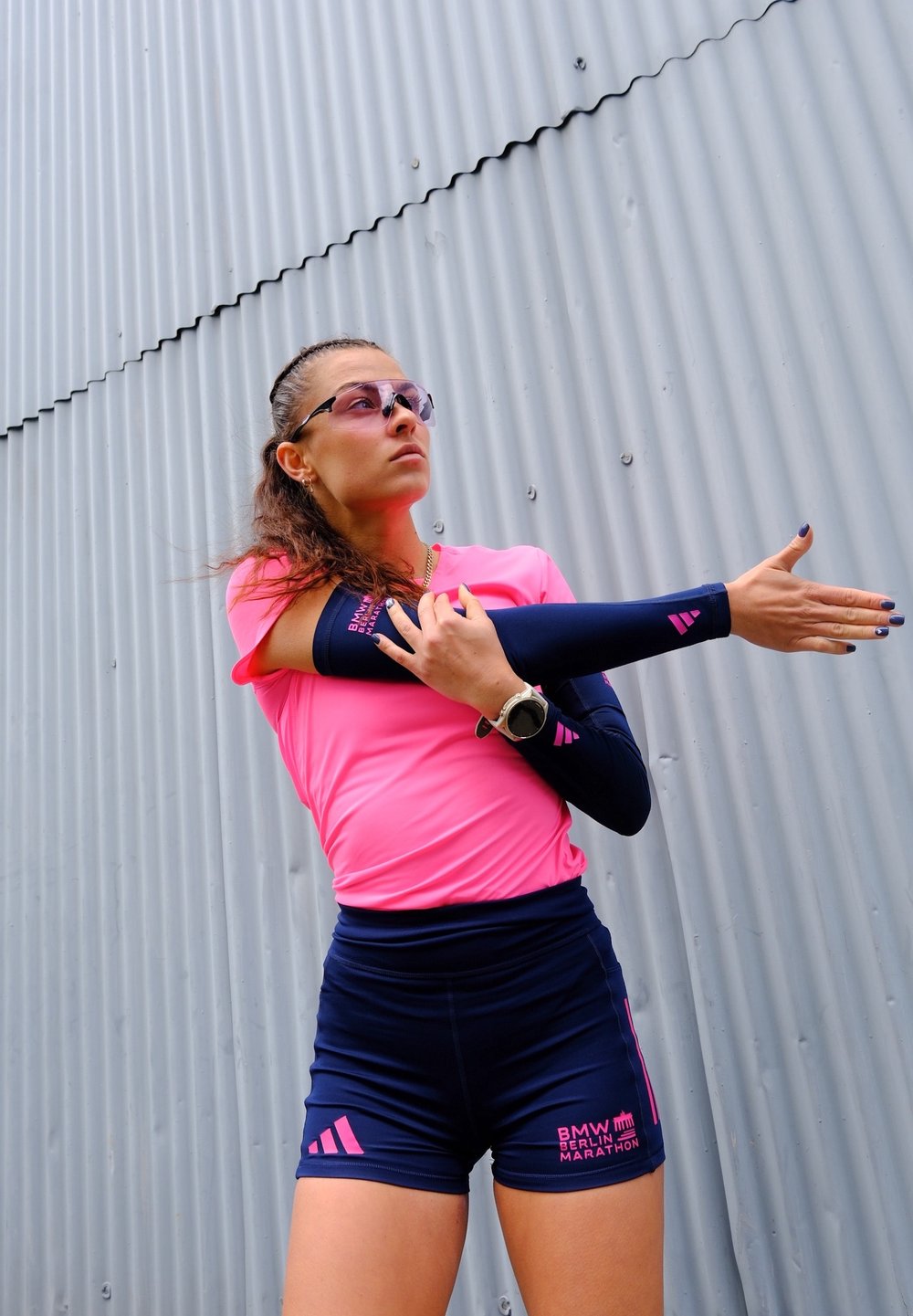 Atleta femminile in abbigliamento sportivo rosa e blu che allunga un braccio contro un muro di metallo ondulato, indossando occhiali da sole e un orologio smartwatch.