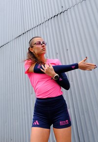 Atleta femminile in abbigliamento sportivo rosa e blu che allunga un braccio contro un muro di metallo ondulato, indossando occhiali da sole e un orologio smartwatch.