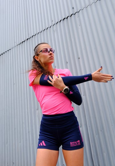 Atleta femenina con ropa deportiva rosa y azul marino estirando su brazo contra una pared de metal acanalado, usando gafas de sol y un reloj inteligente.
