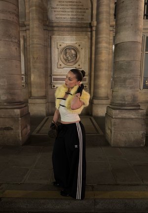 Mujer con estola de piel amarilla, top corto blanco y pantalones Adidas negros, de pie entre columnas de piedra en un pasillo de un edificio histórico por la noche.