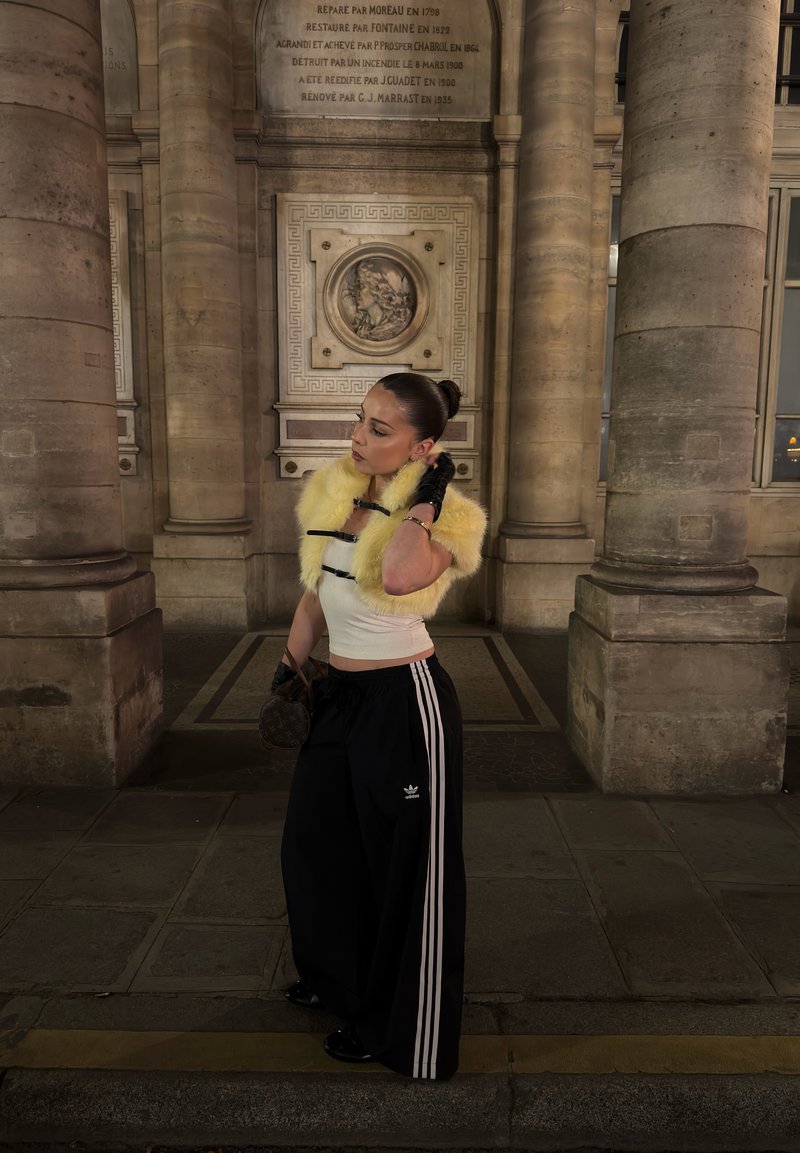 Woman in yellow fur stole, white crop top, and black Adidas pants standing between stone columns in historic building corridor at night.