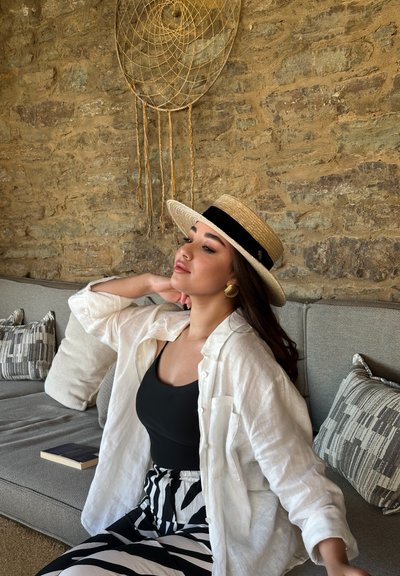 Sombrero de paja de ala ancha con banda negra, camisa de lino blanca, camiseta negra y pantalones a rayas en blanco y negro. Sofá con cojines estampados al fondo.