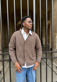 Brown knitted cardigan featuring a V-neck and ribbed cuffs, worn over a white collared shirt. The person is accessorised with rings.