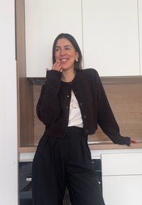 Γυναίκα που χαμογελά και αγγίζει το πρόσωπό της, φορώντας μαύρη ζακέτα, λευκή δαντελένια μπλούζα και μαύρο παντελόνι, στέκεται σε μια μοντέρνα κουζίνα.