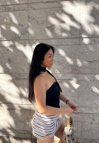 Top negro con cuello halter, falda a rayas con una abertura lateral y un bolso con flecos. Piel suave, cabello largo y oscuro, de pie contra una pared de mármol texturizada.