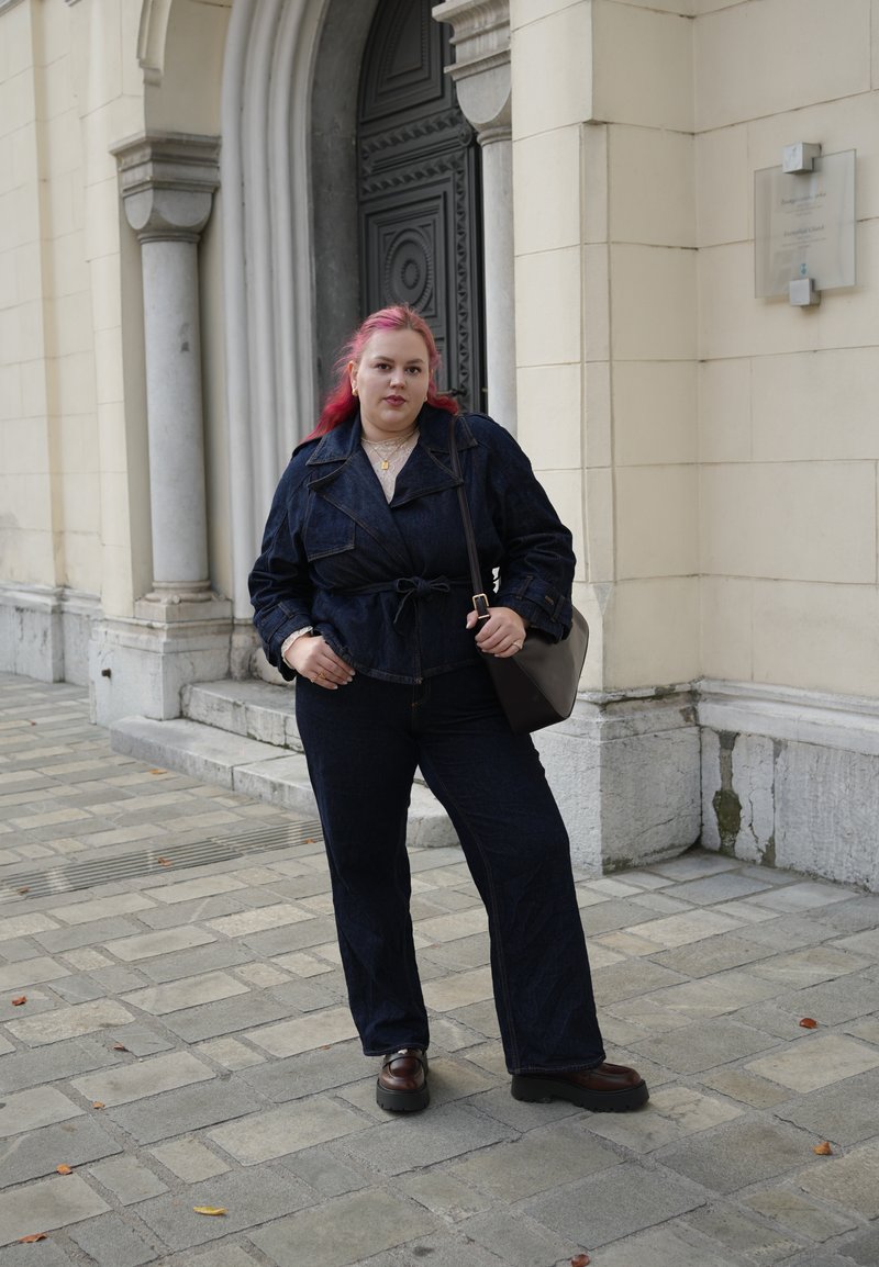 Veste en denim foncé avec une ceinture à nouer à la taille, associée à un jean flare assorti. Un haut gris en dessous et des chaussures marron massives complètent le look.