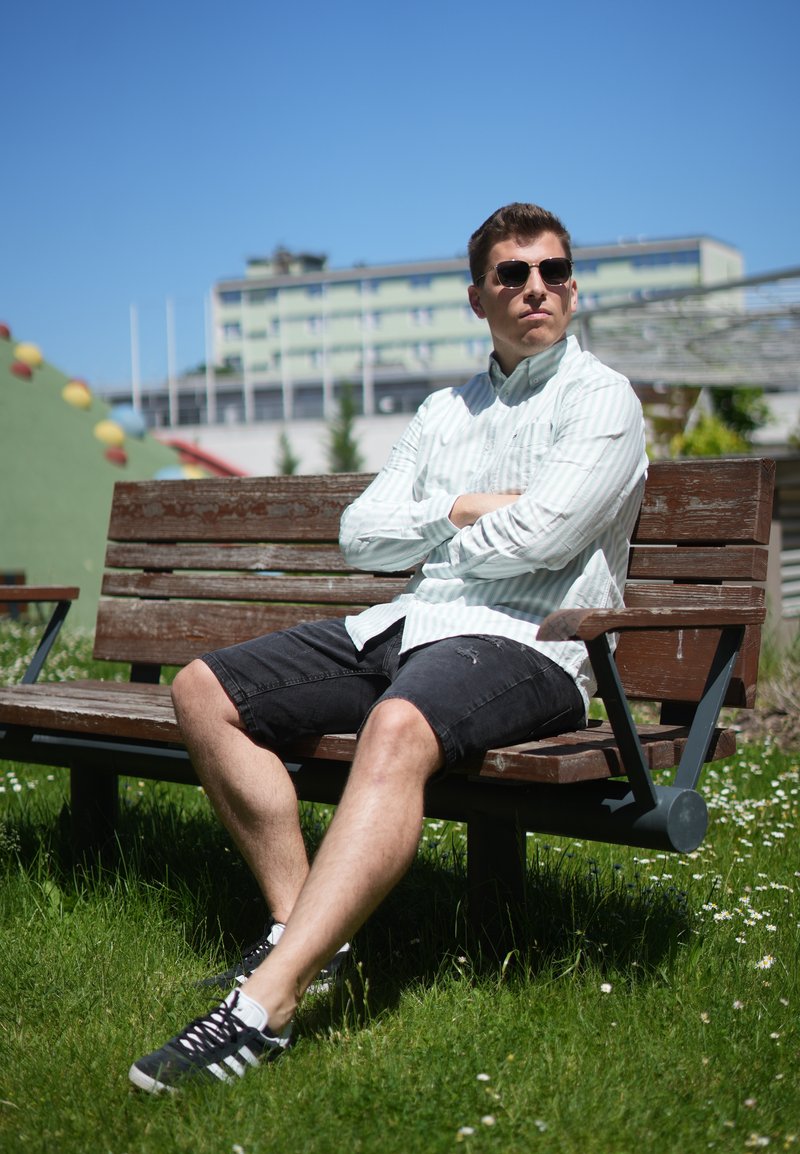 Junger Mann mit Sonnenbrille, weissem Hemd und schwarzen Shorts sitzt auf einer Holzbank in einem sonnigen Park mit Gebäuden im Hintergrund.