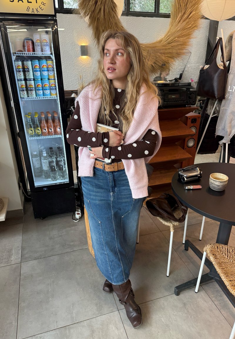 Femme aux longs cheveux blonds portant un haut à pois, un pull rose, une jupe en jean et des bottes marron, tenant un livre dans un café chaleureux.