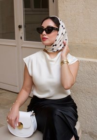 Weißes Satinoberteil mit kurzen Ärmeln, schwarzer Satinrock, cremefarbene Handtasche mit goldenen Details und weißes Tuch mit schwarzen Punkten. Schwarze Sonnenbrille.