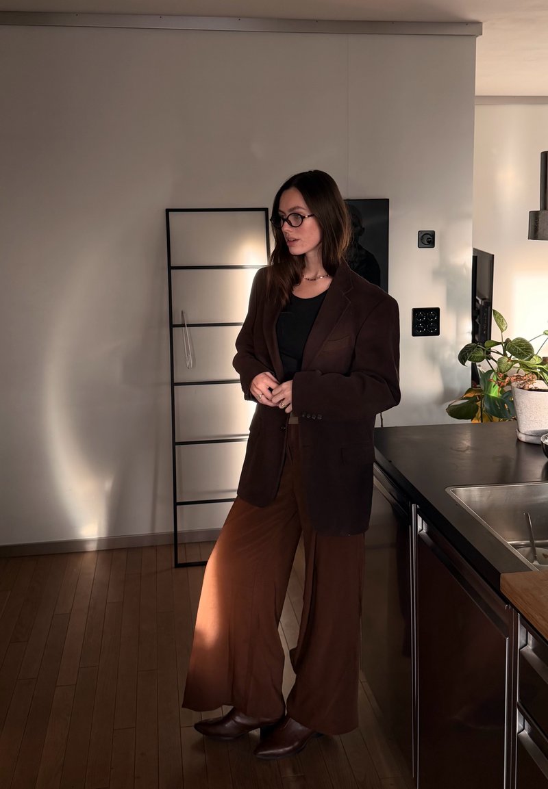 Femme portant un blazer marron et un pantalon à jambes larges, debout dans une cuisine moderne à côté d'un comptoir avec des plantes et un évier.