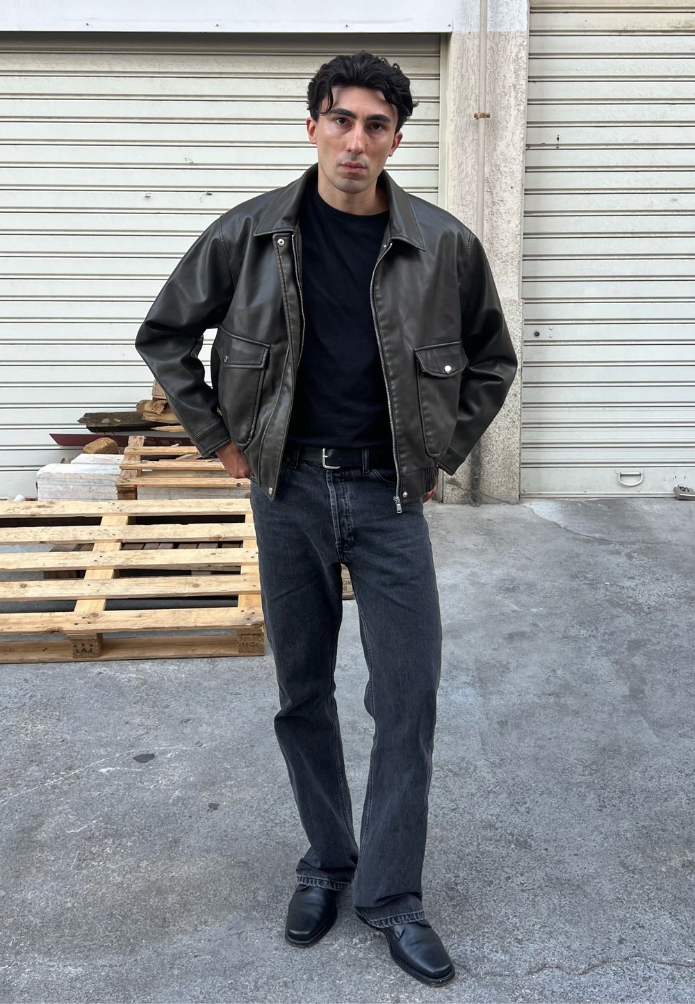 Veste en cuir vert foncé avec poches latérales, associée à un t-shirt noir et un jean noir délavé, le tout complété par des chaussures noires élégantes.
