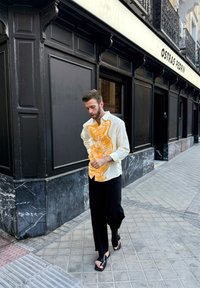 Viegls, strukturēts krekls ar oranžu grafiku, apvienots ar brīviem melniem biksēm un melniem sandaliem, uz pilsētas trotuāra fona.