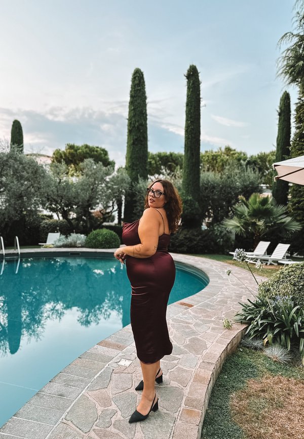 Burgundy satin slip dress with a fitted silhouette, knee-length hem, paired with black pointed heels, against a poolside background.