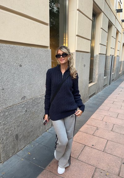 Suéter de punto trenzado azul marino con cuello de cremallera, combinado con pantalones de chándal gris claro con franjas laterales negras, y zapatillas blancas.