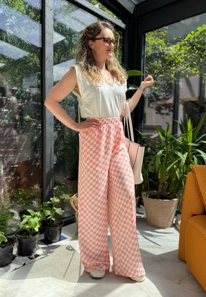 Top blanco de cuello en V combinado con pantalones de pierna ancha a cuadros rosas. Tejido ligero, cinturilla elástica y zapatillas. Entorno interior con plantas.
