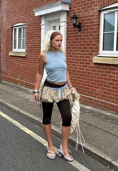 Top sin mangas de color azul claro, pantalones cortos de cuadros holgados con un acento azul, leggings negros y sandalias de tacón alto plateadas. Lleva una bolsa blanca.