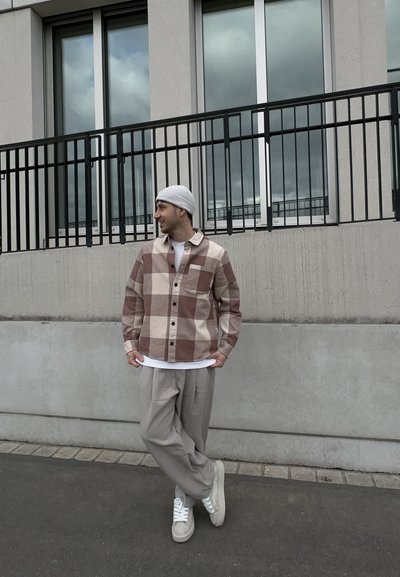 Hombre con gorro blanco, camisa a cuadros marrón y beige, pantalones beige y zapatillas blancas, de pie contra una pared de concreto y una barandilla.
