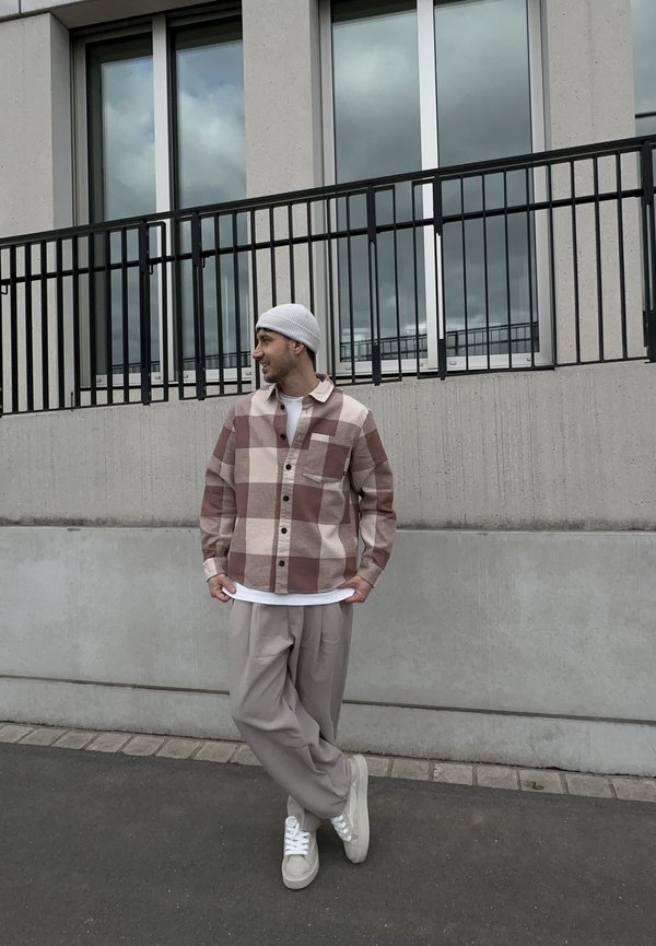 Homme portant un bonnet blanc, une chemise à carreaux marron et beige, un pantalon beige et des baskets blanches, debout contre un mur en béton et une rambarde.