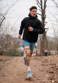 Corredor com uma jaqueta preta com capuz e calções curtos azul claro, com tatuagens nas pernas, usando sapatilhas de corrida azuis em um caminho de terra entre árvores despidas.