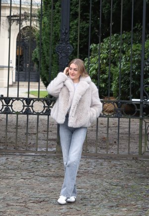 Abrigo de piel sintética en beige claro, con un cuello ancho y cierre frontal, combinado con jeans de campana azul claro y zapatillas blancas. De pie junto a una puerta.