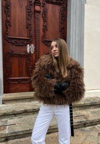 Jeune femme aux longs cheveux portant une veste marron duveteuse et un pantalon blanc, debout les bras croisés sur des marches en pierre devant une porte en bois ornée.