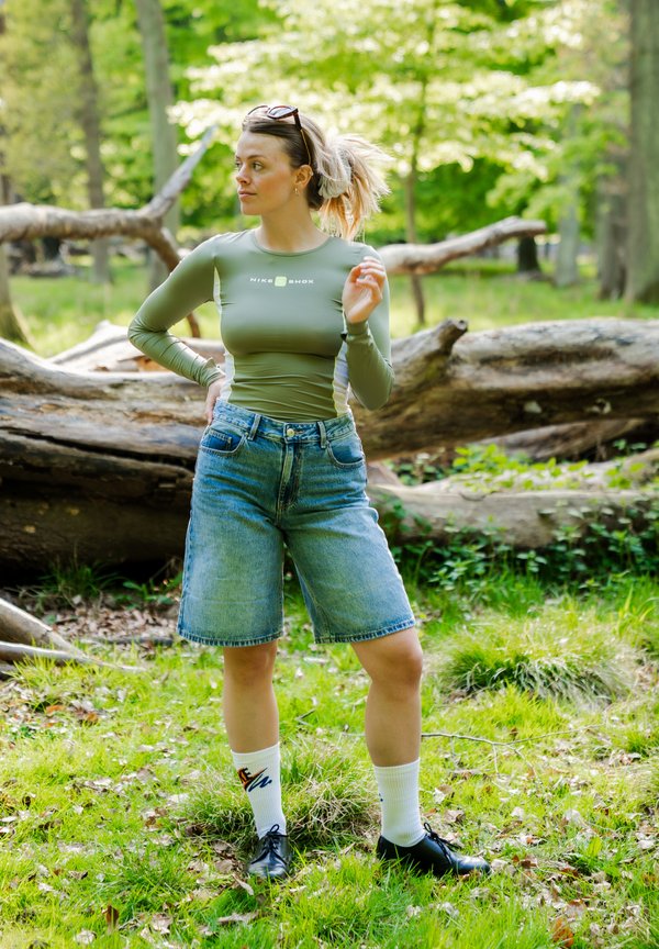 Femme en chemise à manches longues verte, shorts en jean et chaussures noires debout sur l'herbe avec des troncs d'arbres tombés en arrière-plan d'une forêt ensoleillée.