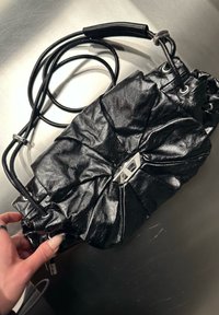 Saco de ombro em couro preto com pregas, fecho prateado e alça ajustável, segurado por uma mão com unhas compridas sobre uma superfície metálica.