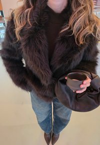 Barna műszőrme kabát, puha, bolyhos textúrával, sötét pulóver fölött viselve. Egyik kézben fekete bőrborítójú kis kézitáska és egy kis csésze található.