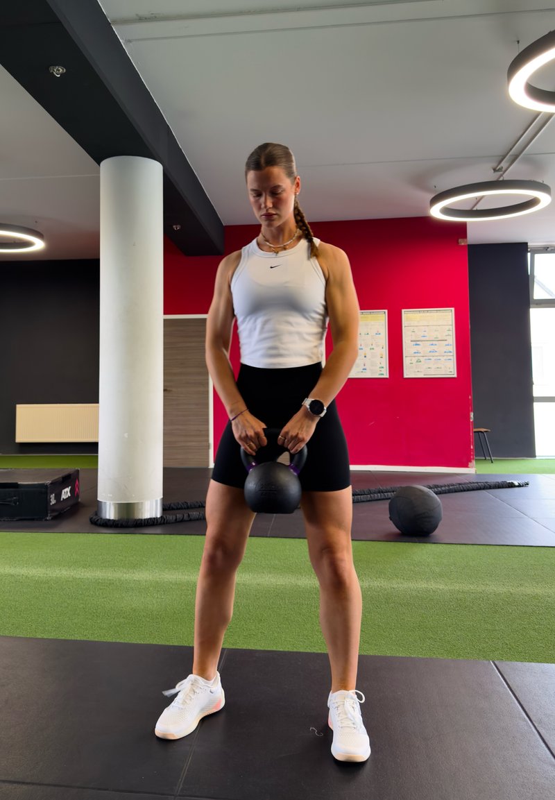 Femme en débardeur blanc et short noir tenant un kettlebell, debout sur le sol de la salle de sport avec du matériel d’exercice et un mur rouge en arrière-plan.
