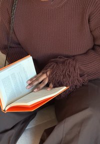 Pull en maille marron avec des manches texturées à franges, associé à un vêtement marron. Une main tient un livre ouvert à la couverture orange et aux pages blanches.