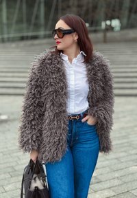 Manteau gris duveteux avec un motif texturé, porté sur une chemise blanche à boutons. Un jean bleu taille haute et un sac à main noir complètent la tenue.